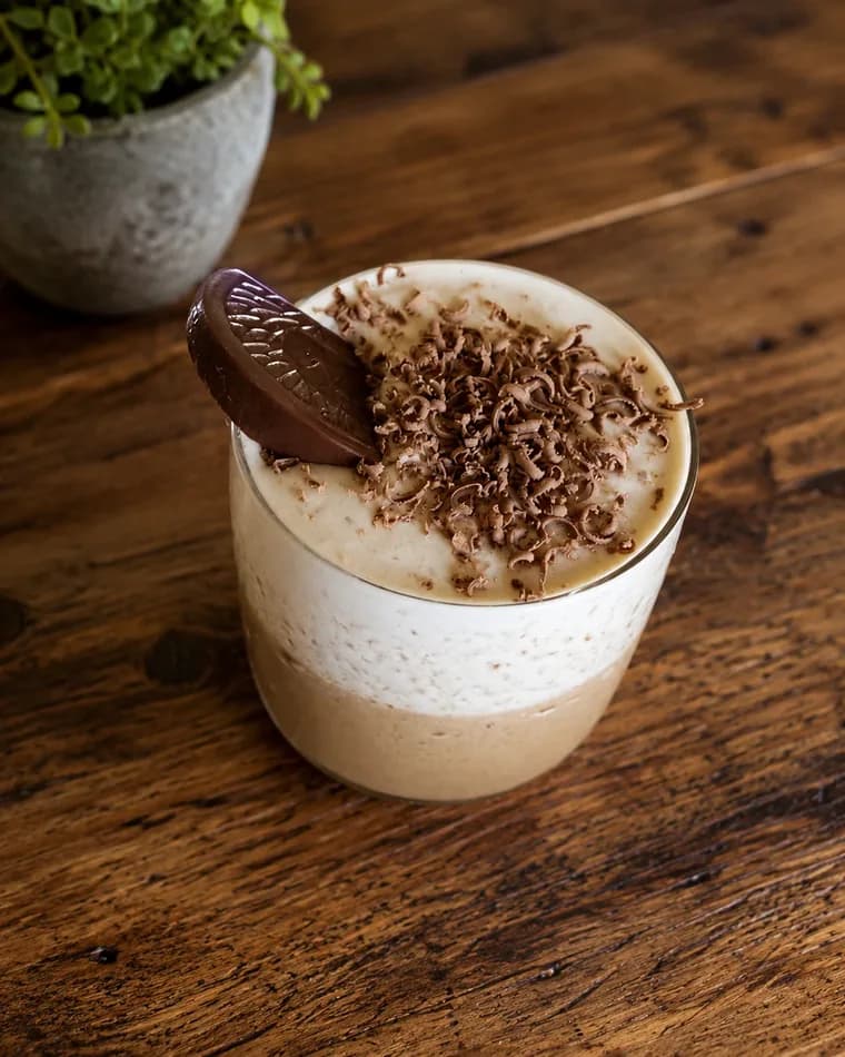 A tumbler of a pale blended drink on a dark wooden table, topped with a thick layer of grated chocolate and a chocolate wafer, beside a small potted plant.